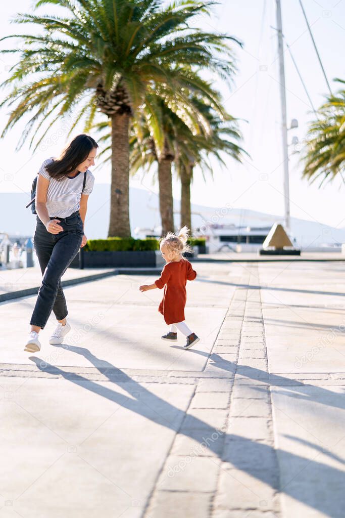 A cute 2-year old is running after her mommy among palm trees on a boat pier
