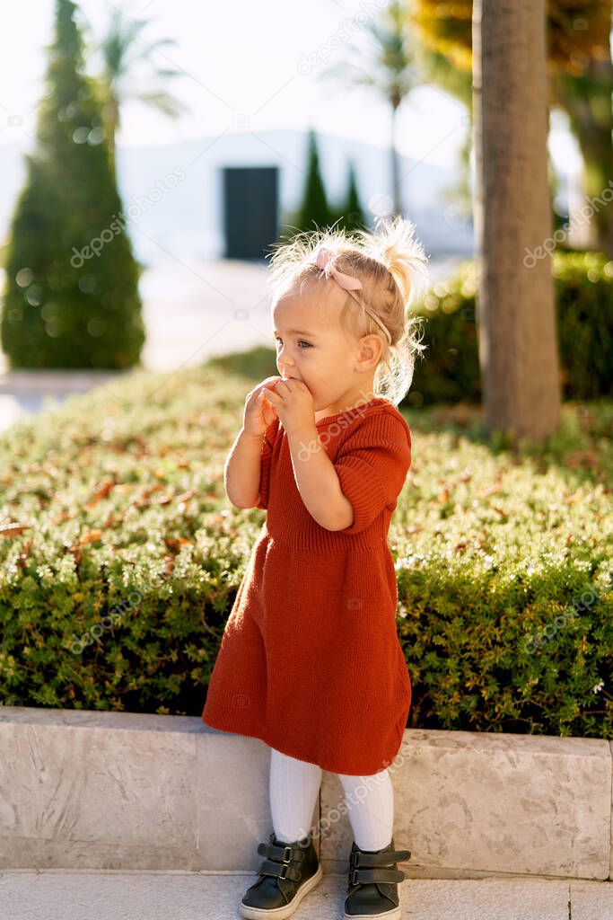 Pretty little girl in a terracotta colored dress and white tights is snacking on a pie in a park on a sunny day