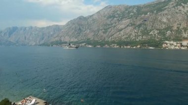 Kotor Körfezi manzarası, güzel dağlar, yelkenli bir gemi ve iskelede güneşlenen insanlar.