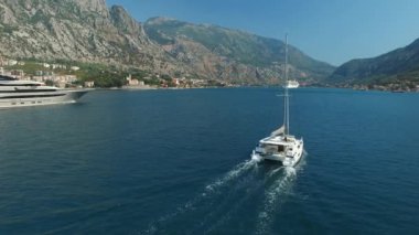 Küçük yelkenli yat ve büyük olan Kotor Körfezi boyunca yelken açıyor. Arkalarında güzel dağlar ve küçük şirin bir kasaba var.