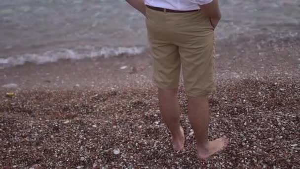 Un homme pieds nus en short se tient sur une plage de galets près de l'eau — Video