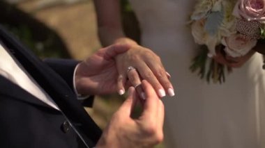 Damat gelinin parmağına yüzük takar, gelin elinde buket tutar.