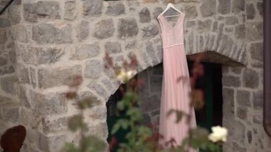 Güzel, soluk, pembe bir gelinlik girişte asılı taş bir duvarda, rahat bir avluda.