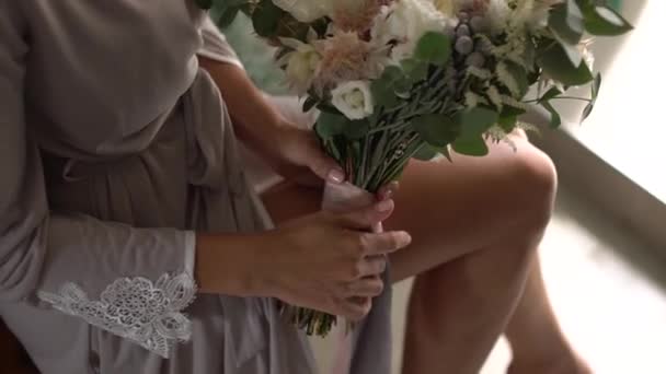 Een vrouw in een zijden badjas houdt een bruidsboeket op schoot en speelt met lange linten — Stockvideo