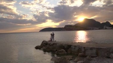 Gelin ve damat, sakin denizin arka planında rıhtımda sarılıyorlar.