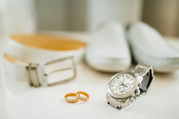 Close-up de alianças de casamento da noiva e do noivo em um fundo branco com um relógio de pulso, cinto branco e mocassins mens. — Fotografia de Stock