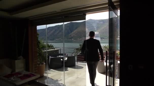 Чоловік у костюмі виходить на балкон готельного номера з прекрасним видом на затоку Котор. — стокове відео