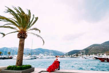 Kırmızı elbiseli bir adam ve bir kadın rıhtımda birbirlerine sarılıyorlar.