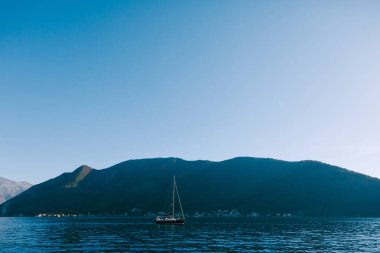 Mavi gökyüzü ve Perast şehrindeki yüksek dağlar. Suyun üzerinde yelken açan bir yat..