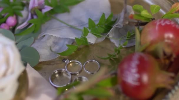 Anillos de boda, granadas jóvenes con nombres escritos en ellos, capullos de rosa y ramitas verdes en una bandeja de plata brillante — Vídeos de Stock