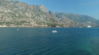 Bir turist teknesi Kotor Körfezi 'nin ortasında yelken açar. Önünde küçük bir kasaba, büyük bir yat ve küçük bir tekne vardır.