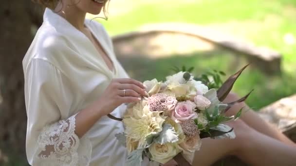 Una chica en un elegante peignoir se sienta en la calle con un ramo de novia en sus manos — Vídeos de Stock