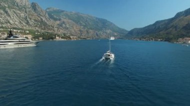 Büyük yat ve küçük yelkenli yat Kotor Körfezi boyunca yol alıyor. Arkalarında güzel dağlar ve küçük şirin bir kasaba var.