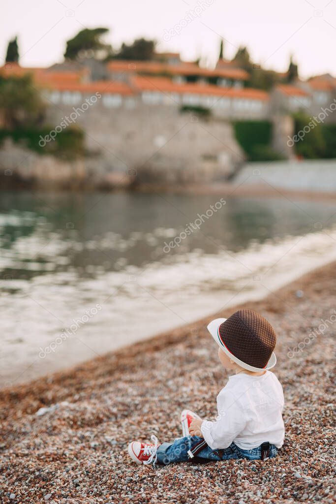 A blue-eyed little boy in a cute hat is sitting on the seashore against the background of the Sveti Stefan island, back view