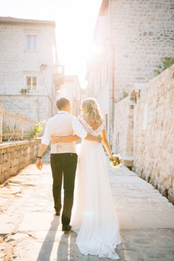 Gelin ve damat Perast caddelerinde birbirlerine sarılıyorlar. Beyaz tuğladan evler, arka manzara.