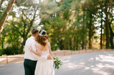 Gelin ve damat parkta yürüyorlar, birbirlerine sarılıyorlar, gelin bir buket tutuyor. 