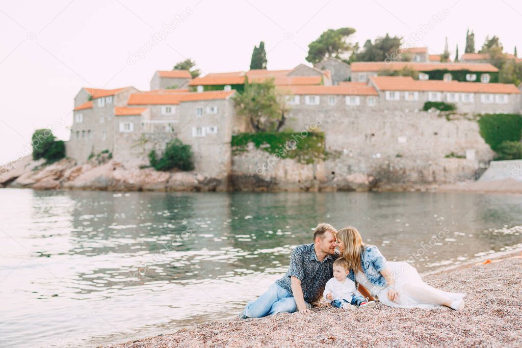 Mom, dad and their little son lying on the seashore against the backdrop of the Sveti Stefan island, parents are kissing