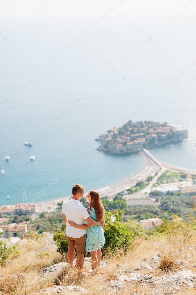 A man and a woman stand embracing on a mountain overlooking the island of Sveti Stefan and look at each other 