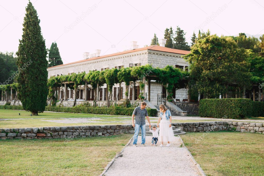 Mom, dad and little son are walking along the path near the beautiful Villa Milocer, parents hold the childs hands 