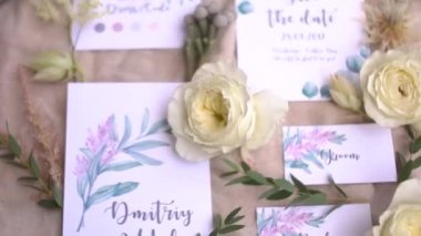 Üzerinde suluboya çizimler, çiçekler ve dallar olan düğün davetiyeleri pastel kumaşın üzerinde.