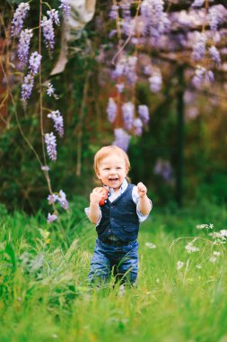 Kızıl saçlı çocuk arka planda Wysteria ağacıyla yeşil çimlerin üzerinde duruyor.