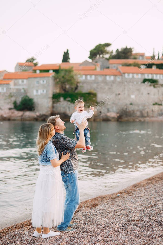 Mom and dad are holding their little son in their arms by the sea against the backdrop of the Sveti Stefan island
