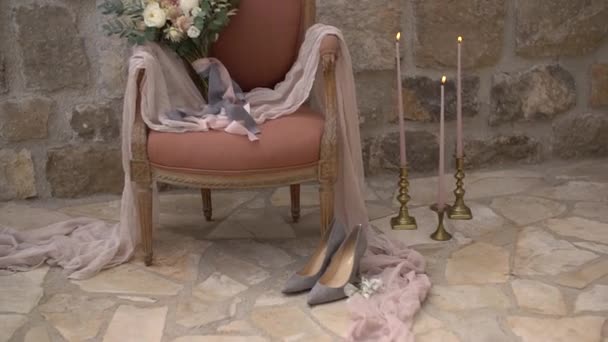 A wedding bouquet in an armchair and delicate fabric, and on the floor are the brides shoes and burning candle — Stock Video