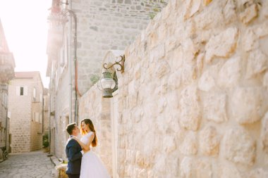 Gelin ve damat, Perast 'in eski güzel bir sokağında, beyaz bir taş duvarın yanında sarılıyorlar. 