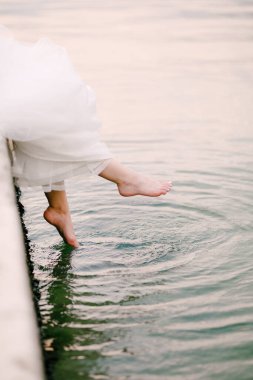 Gelin iskelede oturur ve çıplak ayaklarıyla suda sallanır. Bacakları gelinliğin altından dışarı bakar. 