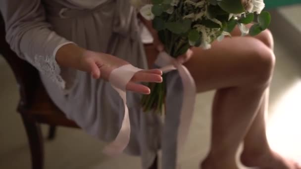 Een vrouw in een zijden badjas houdt een bruidsboeket op schoot en speelt met lange linten — Stockvideo