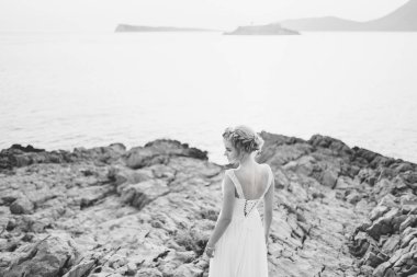 Deniz kıyısındaki bir kayanın üzerinde narin bir gelin duruyor. Önünde bir ada, arkası manzaralı, siyah beyaz bir fotoğraf.