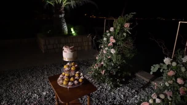 Stojan s košíčky a dort se smetanovými růžemi na dřevěném stole v blízkosti svatebního oblouku — Stock video