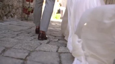 Damat ve gelinin bacakları kaldırımlı yolda yürüyor, gelinlerin etekleri rüzgarda sallanıyor. 
