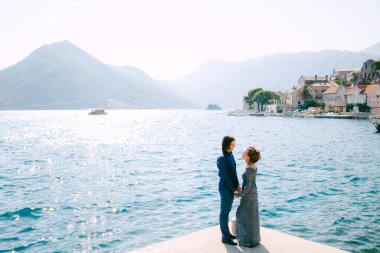 Modaya uygun gri gelinlikli gelin ve damat eski Perast kasabasının deniz kıyısında el ele tutuşuyorlar.