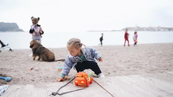 Little girl plays in the sand on the beach. Two-year-old child in a denim jacket playing with plastic toys on a sandy beach in spring. — Stock Video