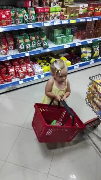 Budva, Czarnogóra - 01 sierpnia 2020: Mała dziewczynka spaceruje z koszykiem w supermarkecie. Dziecko pomaga robić zakupy w sklepie. Roczna córka nosi czerwony plastikowy koszyk ze sklepu.. — Wideo stockowe