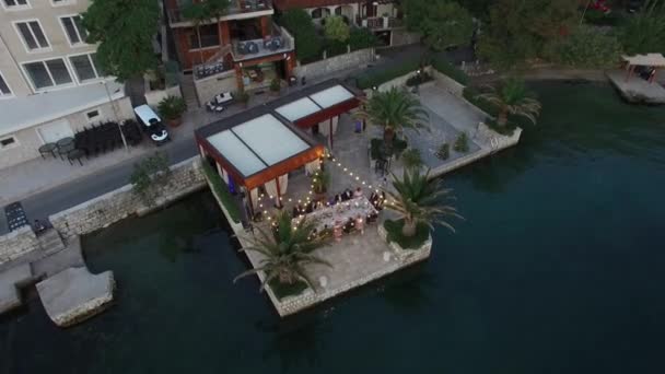 Уютный ресторан у моря с блестящими гирляндами и свадебным банкетом на пирсе в Которском заливе, вид сверху — стоковое видео