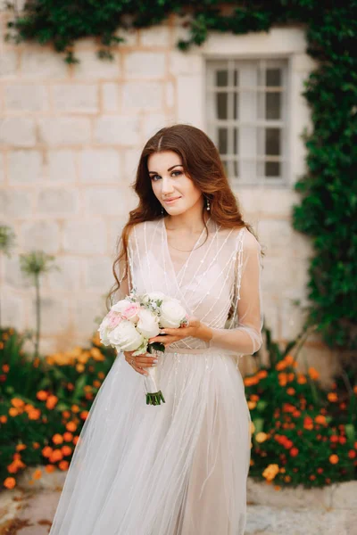 Egy menyasszony csokorral a kezében áll egy ház falánál, zöld liánával és narancsvirágokkal Perastban. — Stock Fotó