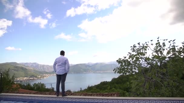 Perast, Montenegro - 10 july 2020: Seorang pria dengan kemeja putih melihat ke kejauhan, melihat ke belakang — Stok Video