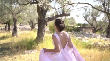 Kotor, Karadağ - 29 Temmuz 2020: Arkası açık, güzel, pembe elbiseli gelin zeytin korusu boyunca koşuyor, arka manzara