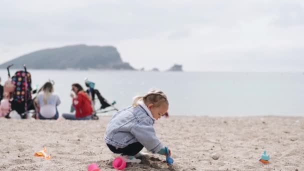 Meisje speelt in het zand op het strand. Twee jaar oud kind in een spijkerjasje spelend met plastic speelgoed op een zandstrand in het voorjaar. — Stockvideo