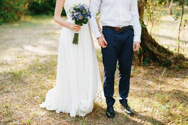 Νύφη με ένα μπουκέτο μπλε λουλούδια και ο γαμπρός στέκονται δίπλα-δίπλα στον ελαιώνα και να κρατήσει τα χέρια — Φωτογραφία Αρχείου