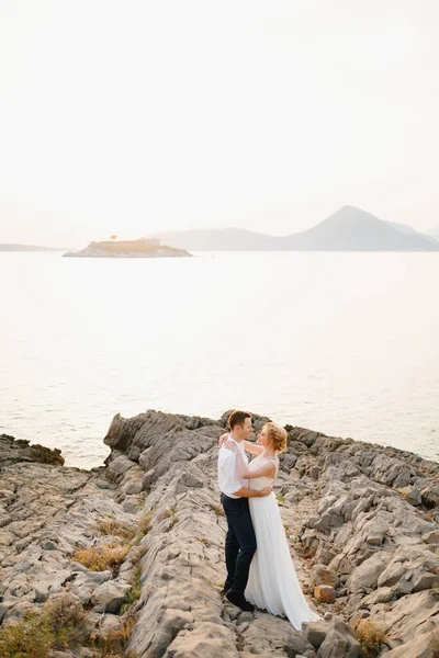 Η νύφη και ο γαμπρός αγκαλιάζονται στα βράχια δίπλα στη θάλασσα με φόντο τα βουνά και το νησί της Μαμούλας — Φωτογραφία Αρχείου