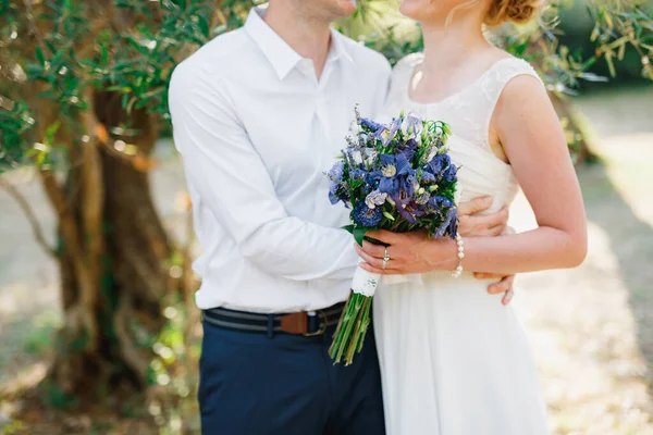 Νύφη με ένα μπουκέτο μπλε λουλούδια και ο γαμπρός αγκαλιάζει τρυφερά στον ελαιώνα — Φωτογραφία Αρχείου