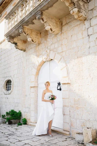 Egy menyasszony egy elegáns ruhában, csokorral a kezében áll egy gyönyörű régi épület fehér ajtajában Perastban. — Stock Fotó