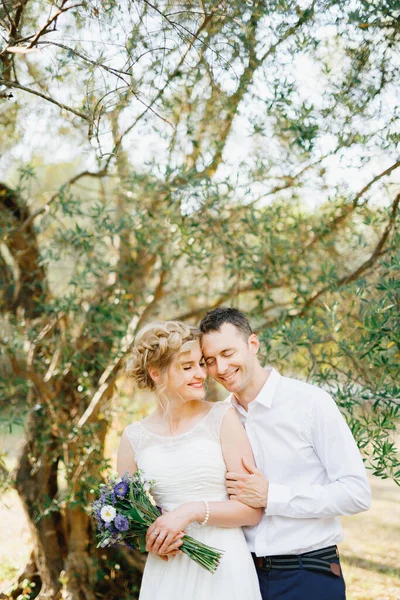 Ο γαμπρός αγκαλιάζει απαλά τη νύφη κοντά στα πράσινα δέντρα στον ελαιώνα, η νύφη κρατά ένα μπουκέτο μπλε λουλούδια — Φωτογραφία Αρχείου