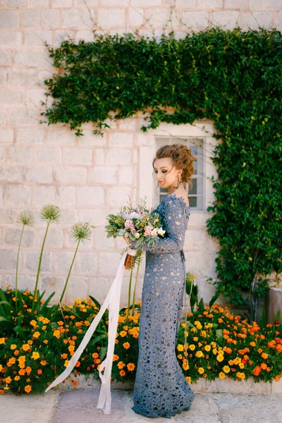 Seorang pengantin dalam gaun biru dengan buket berdiri di dinding rumah dengan liana hijau dan bunga oranye, close-up — Stok Foto