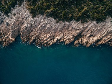Top view of the coastline with cliff, turquoise water, rocks, foam, waves and trees.