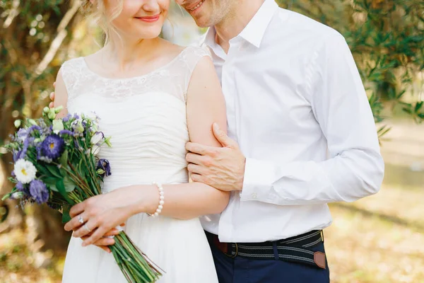 Ο γαμπρός αγκαλιάζει απαλά τη νύφη κοντά στα πράσινα δέντρα στον ελαιώνα, η νύφη κρατά ένα μπουκέτο μπλε λουλούδια — Φωτογραφία Αρχείου
