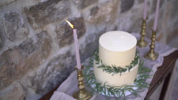 White cream cake, decorated with olive and eucalyptus sprigs, next to the burning candles in golden candlesticks — Stock Video